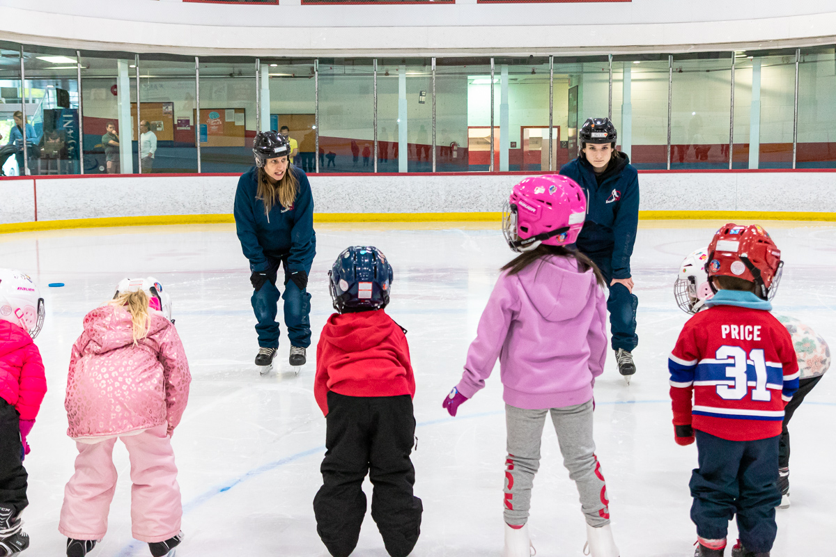 Deux instructrices vêtus en manteaux et pantalons bleus portant des patins apprennent à un groupe d'enfants âgés d'environ 4 ans comment patiner dans une patinoire